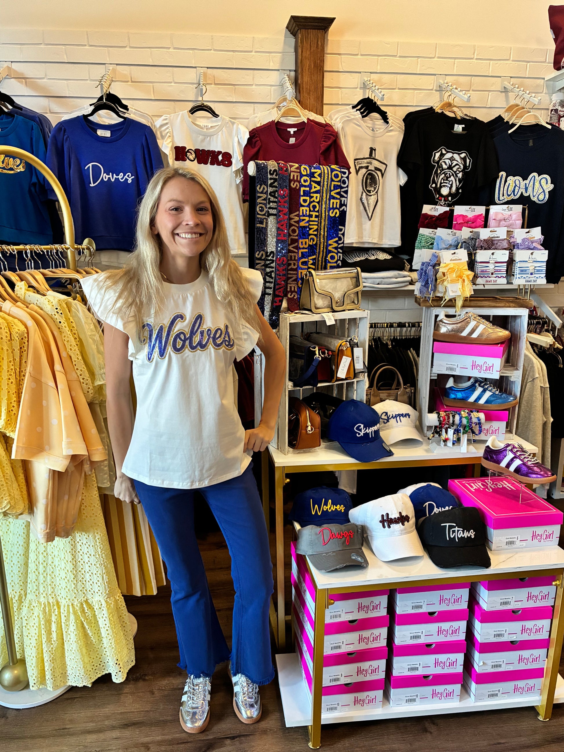 A woman stands in a retail store wearing a white graphic T-shirt with 'Wolves' printed across the chest in blue and gold, paired with blue jeans and silver sneakers. 