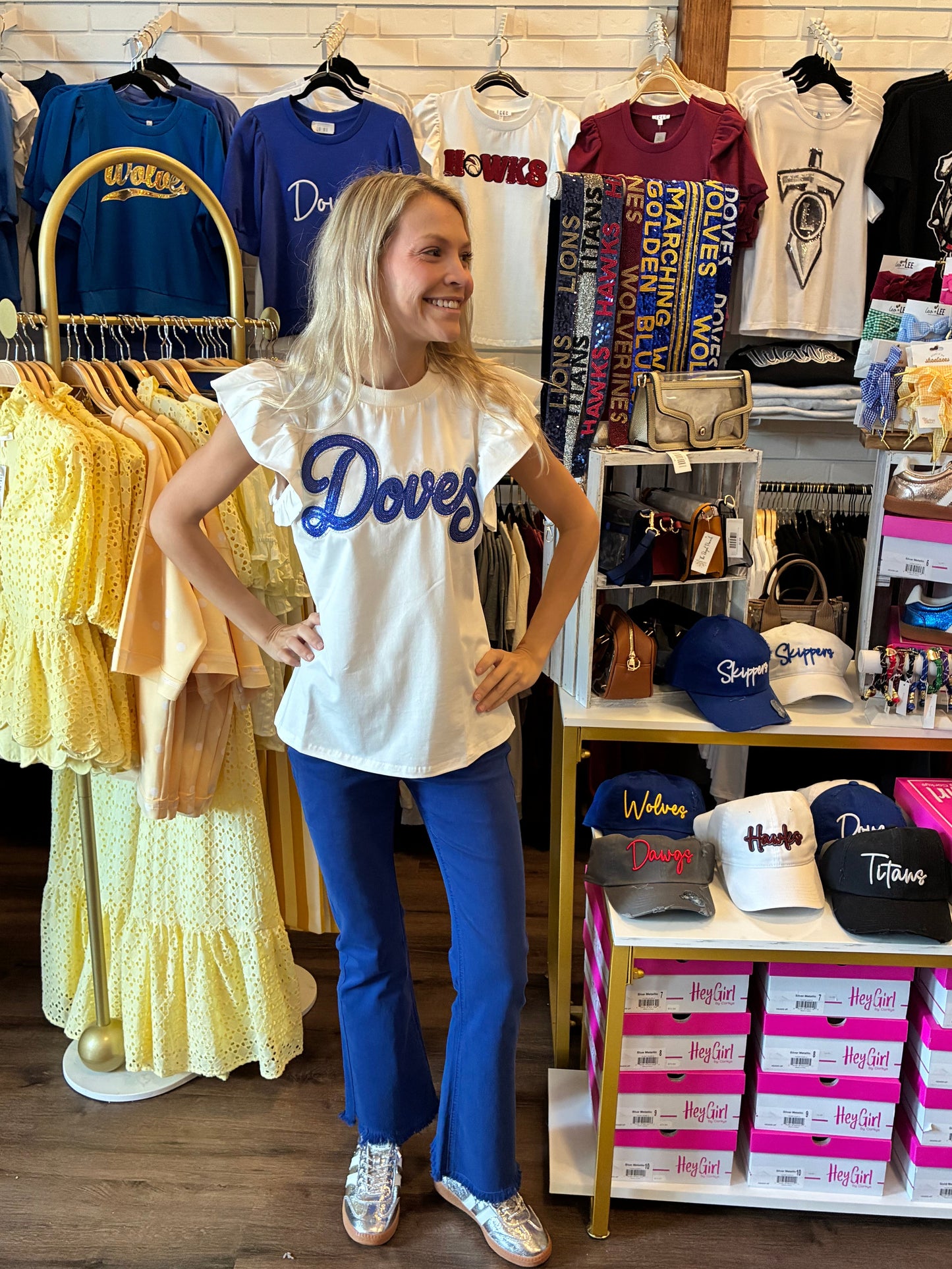 A woman stands inside a retail store wearing a white ruffled short-sleeve t-shirt with 'Doves' printed in blue script across the chest, paired with blue flared pants and white sneakers. The background is filled other 'Doves' branded items.
