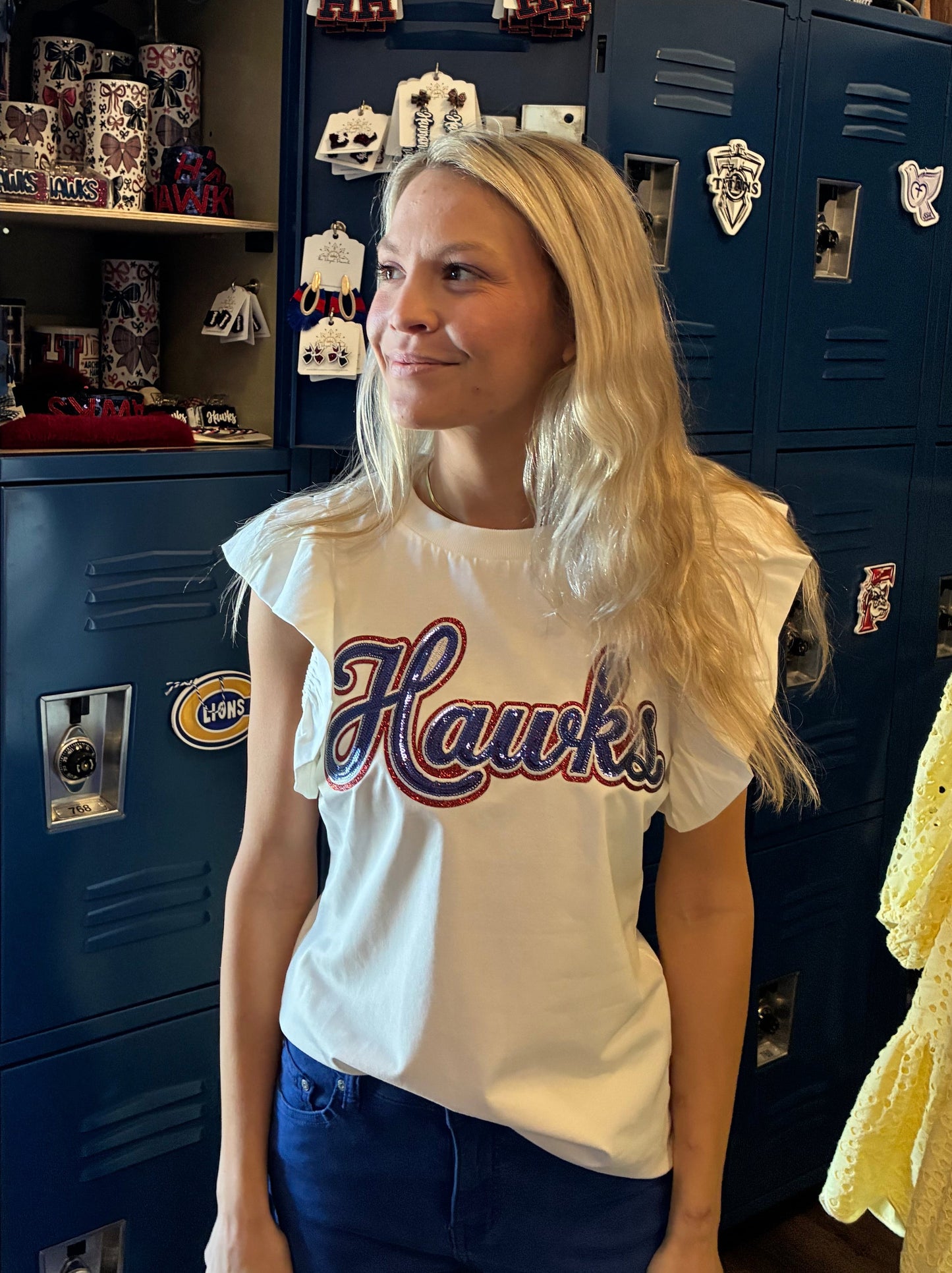 A blonde woman wearing a white graphic t-shirt with 'Hawks' printed in crimson and navy varsity lettering, standing in front of a set of dark blue school lockers decorated with various Hawks Spirit items
