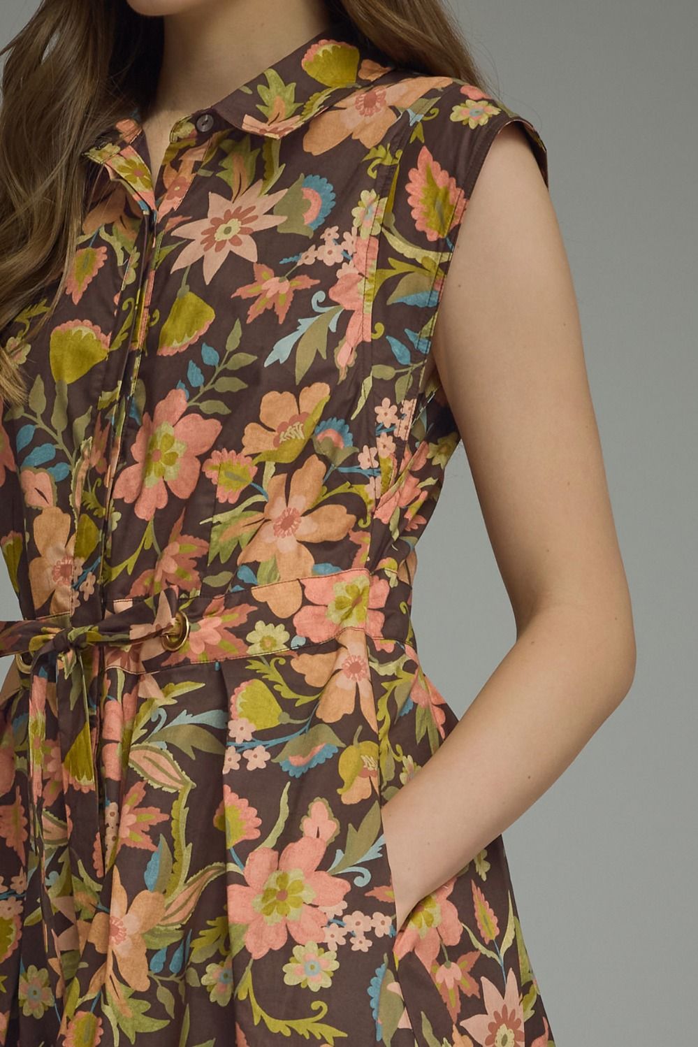 A close-up of a woman wearing a sleeveless collared dress with a brown base and a colorful floral print in pink, orange, green, and blue, featuring a tie waist and side pockets.