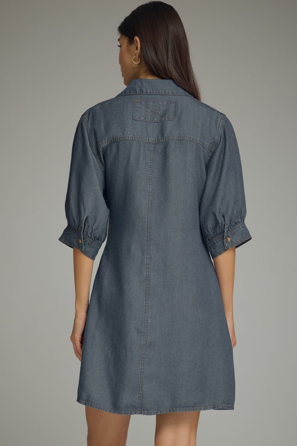 A woman shown from the back wearing a slate blue denim shirt dress with three-quarter sleeves and button cuffs, set against a plain gray studio background.