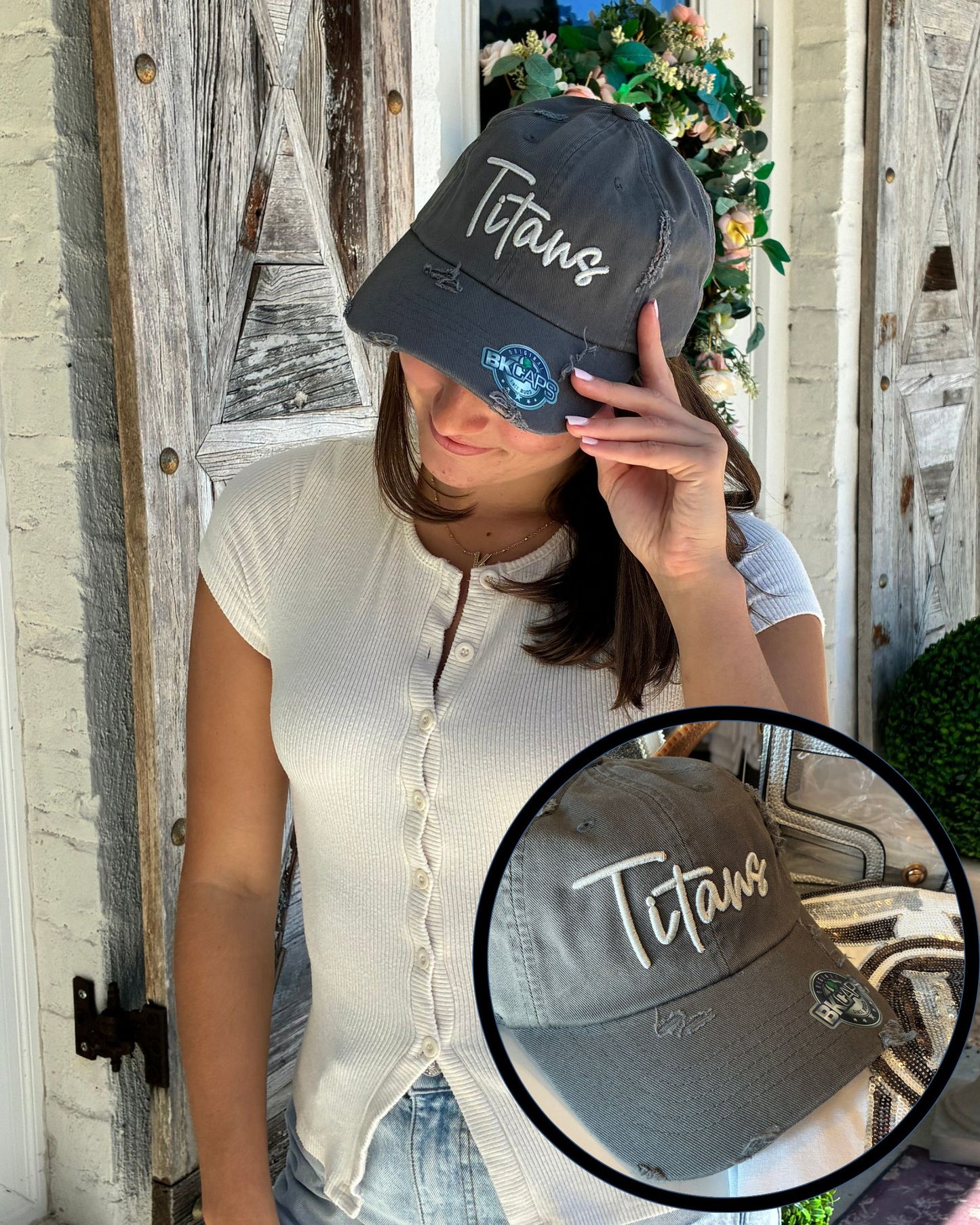 A woman wearing a distressed dark gray baseball cap with 'Titans' embroidered in silver grey script on the front, posed in front of a rustic wooden door. A circular inset shows a close-up of the cap's front panel.