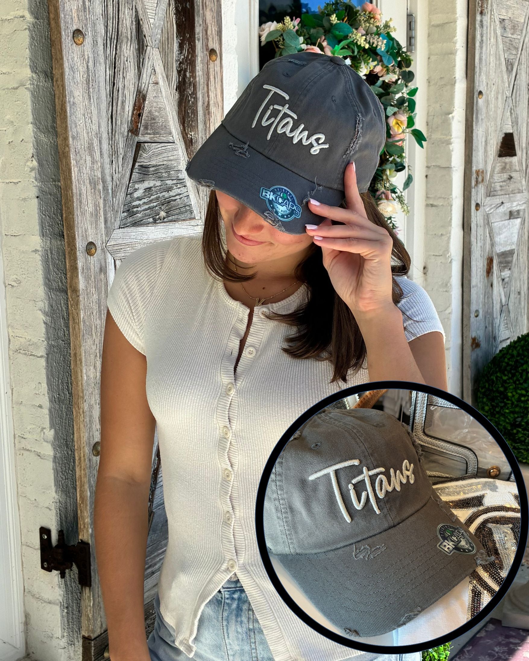 A woman wearing a distressed dark gray baseball cap with 'Titans' embroidered in silver grey script on the front, posed in front of a rustic wooden door. A circular inset shows a close-up of the cap's front panel.