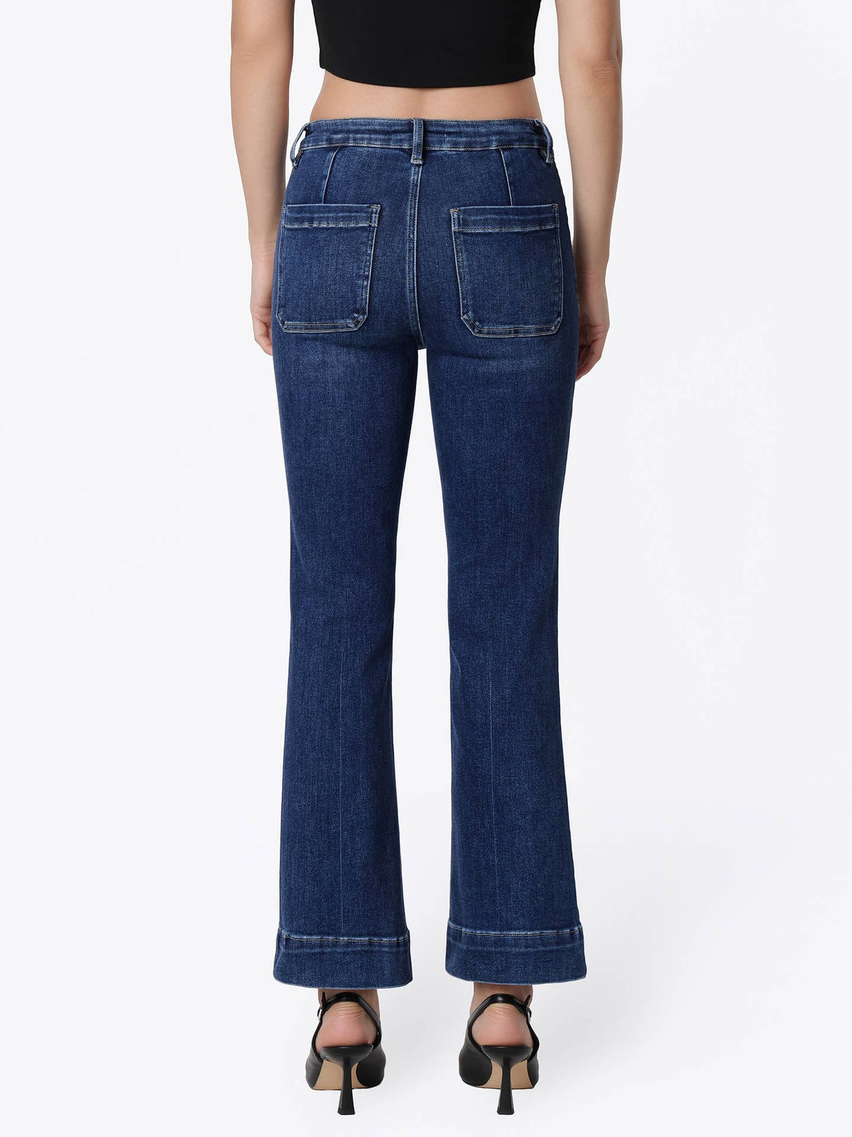 Rear view of a model wearing dark blue flare jeans with finished hem and black high-heeled sandals against a plain white background.