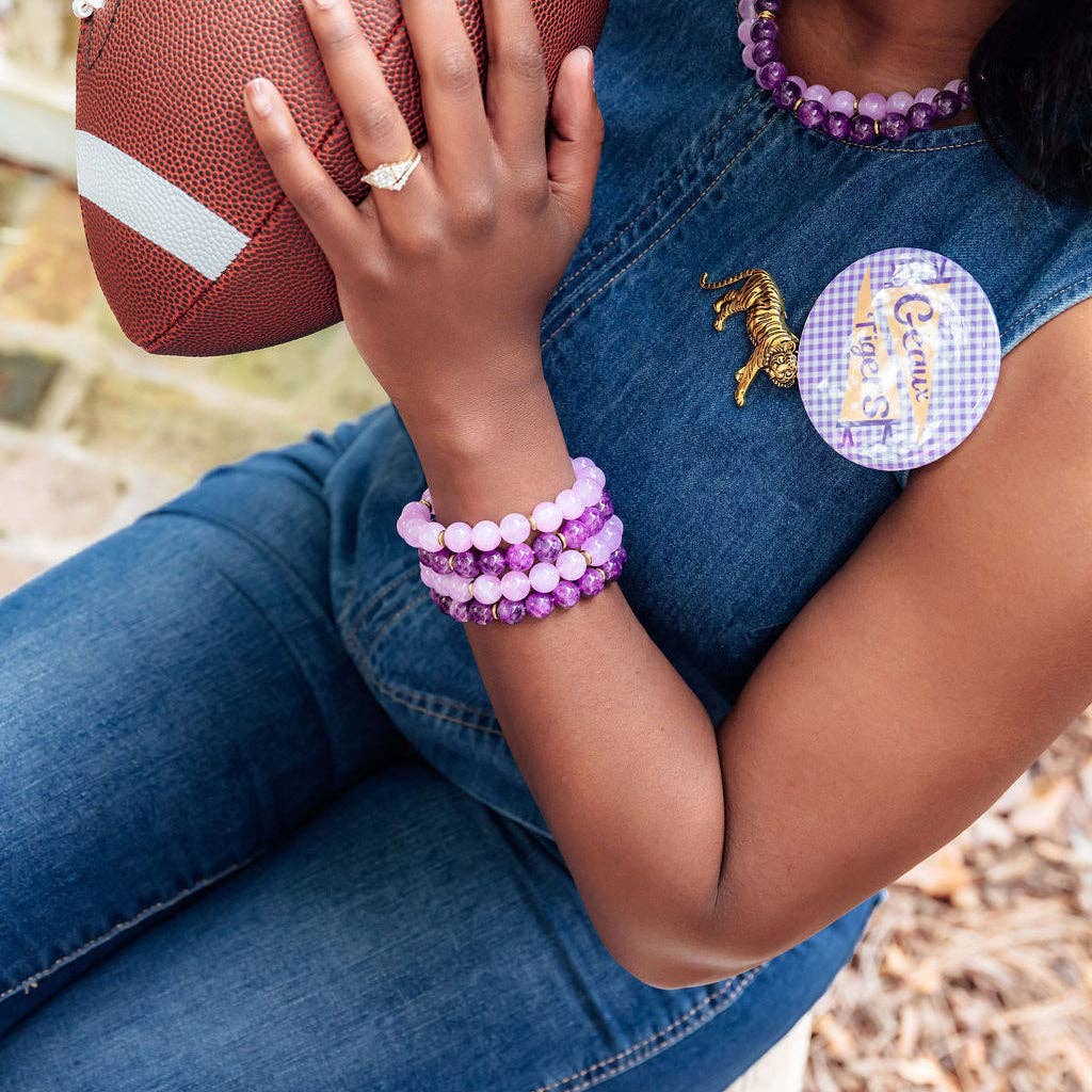 A close-up lifestyle shot of a person wearing purple and pink beaded bracelets on one wrist, a gold horse pin, and a purple gingham patch on a denim outfit, while holding a brown football.