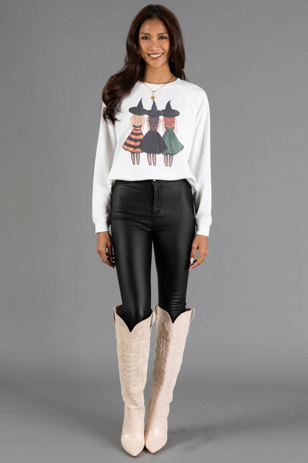 A woman modeling a white crewneck sweatshirt with a graphic of three witch girls, paired with black faux-leather leggings and beige cowboy boots, against a solid gray studio background.