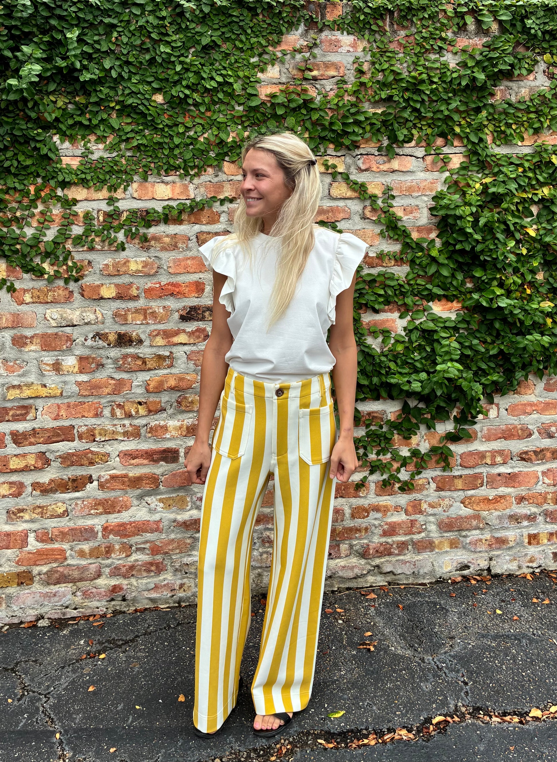 A woman stands outdoors in front of a brick wall covered in green ivy, wearing a white ruffled short-sleeve top and high-waisted yellow-and-white vertically striped wide-leg pants.