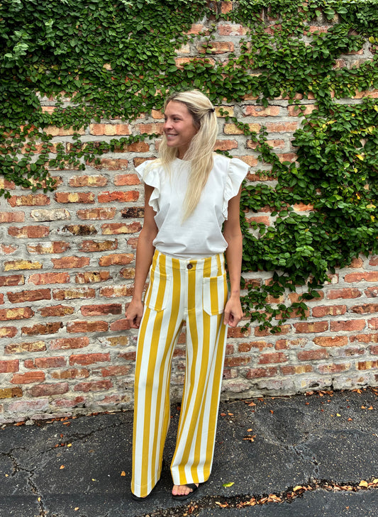 A woman stands outdoors in front of a brick wall covered in green ivy, wearing a white ruffled short-sleeve top and high-waisted yellow-and-white vertically striped wide-leg pants.
