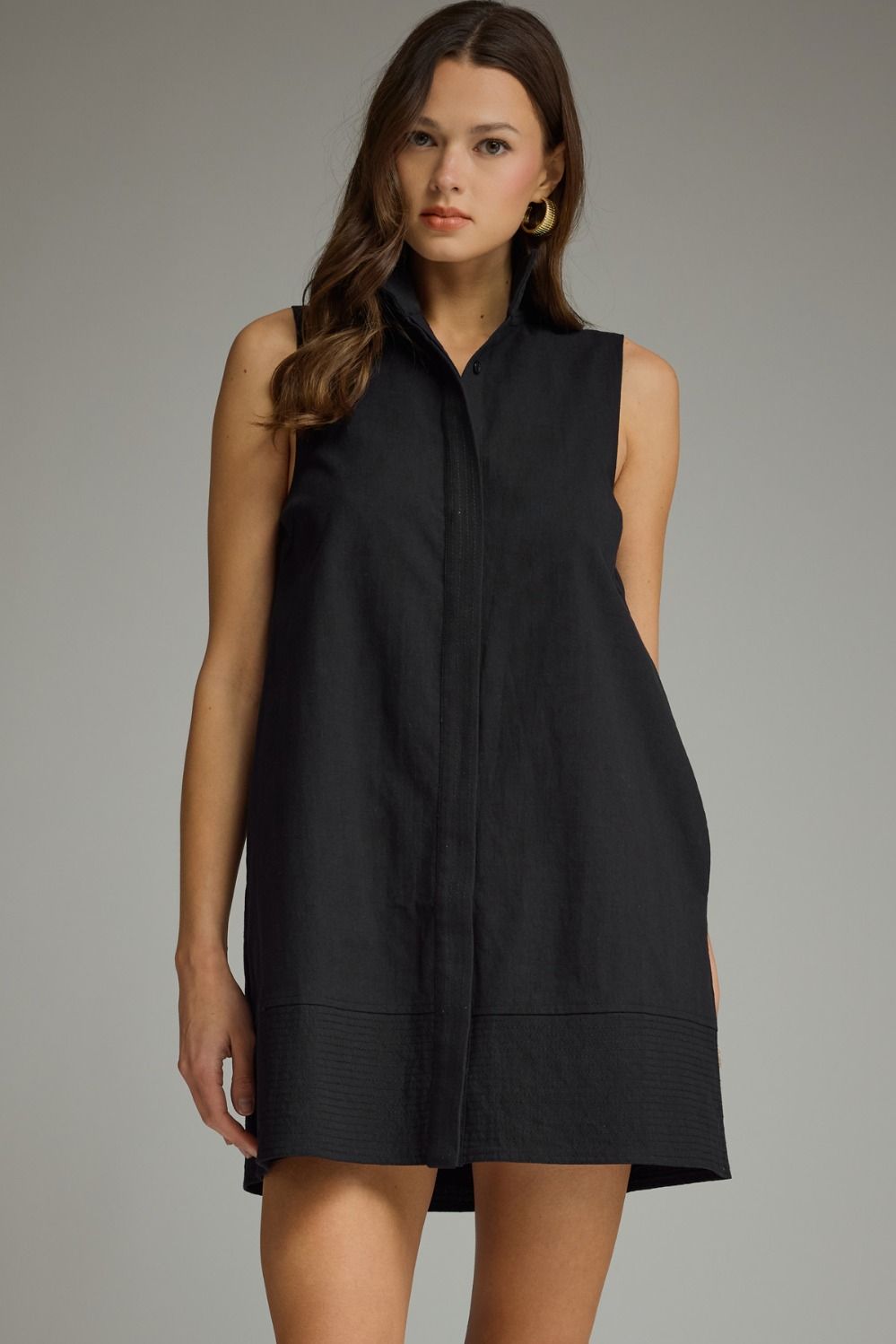 A woman modeling a sleeveless black shift dress with a band collar and front button placket, photographed against a neutral gray studio background.
