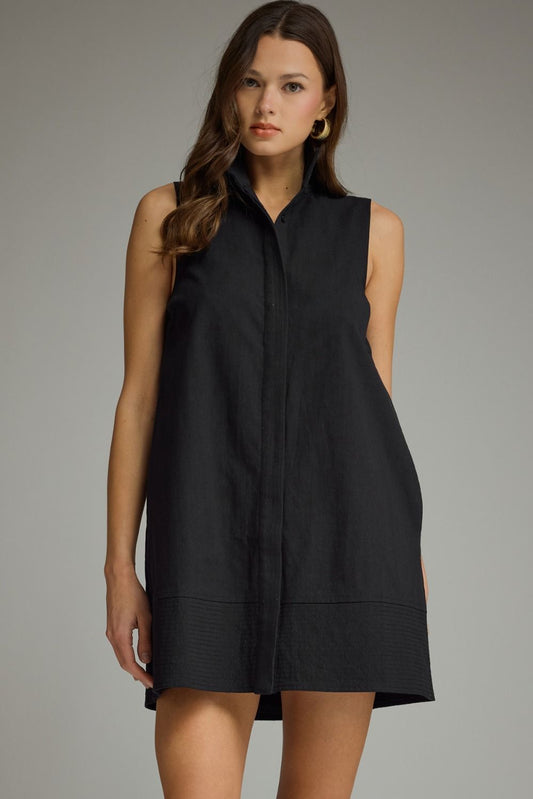 A woman modeling a sleeveless black shift dress with a band collar and front button placket, photographed against a neutral gray studio background.