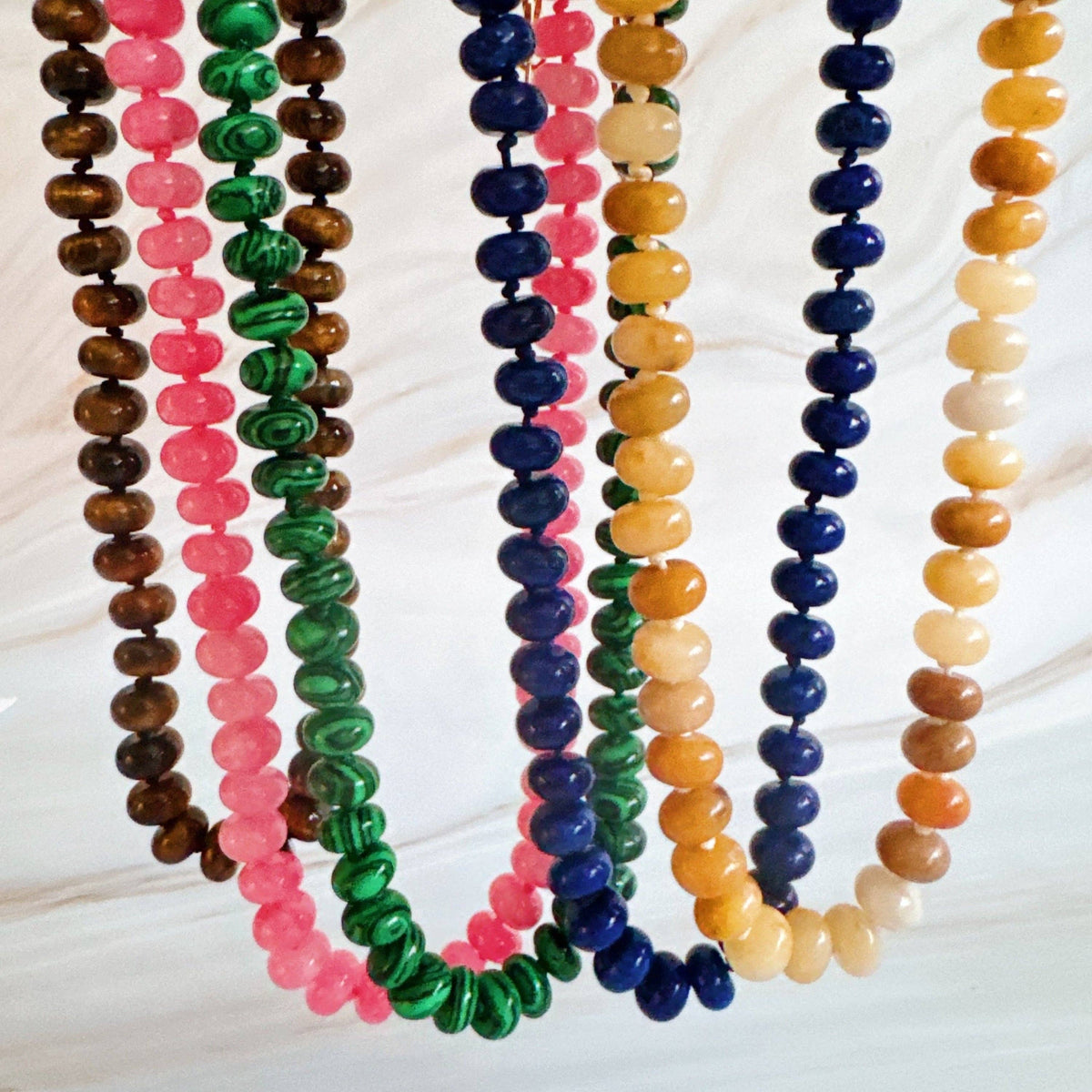 A set of colorful beaded necklaces arranged in vertical strands on a light marble-like surface. The necklaces feature round beads in brown, pink, green, navy blue, yellow, and cream.