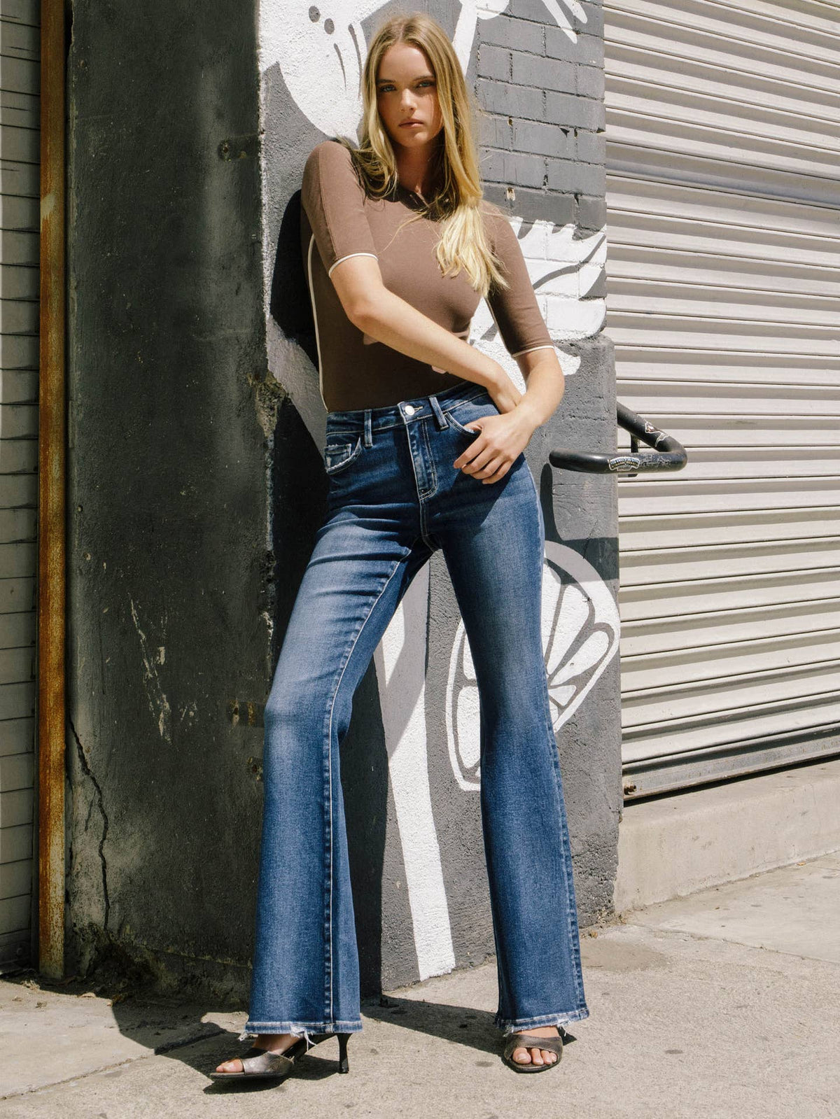 A woman stands outdoors against a painted concrete wall and metal roll-up door, wearing a fitted taupe-brown short-sleeve top and blue flare jeans with dark heeled sandals.