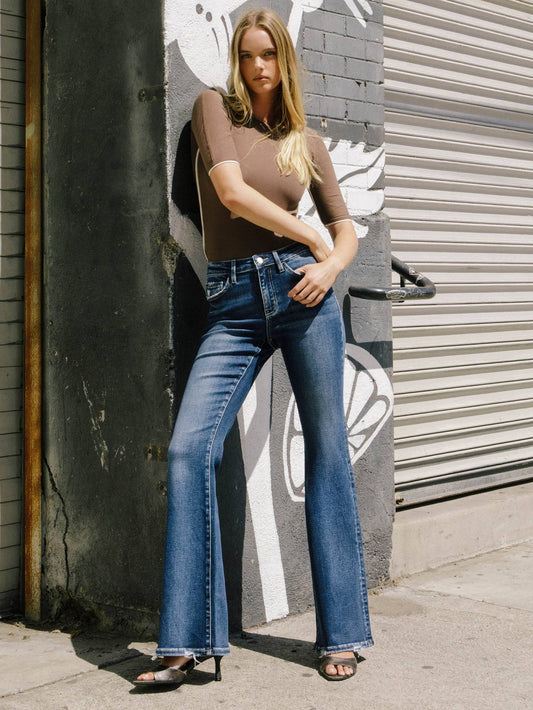 A woman stands outdoors against a painted concrete wall and metal roll-up door, wearing a fitted taupe-brown short-sleeve top and blue flare jeans with dark heeled sandals.