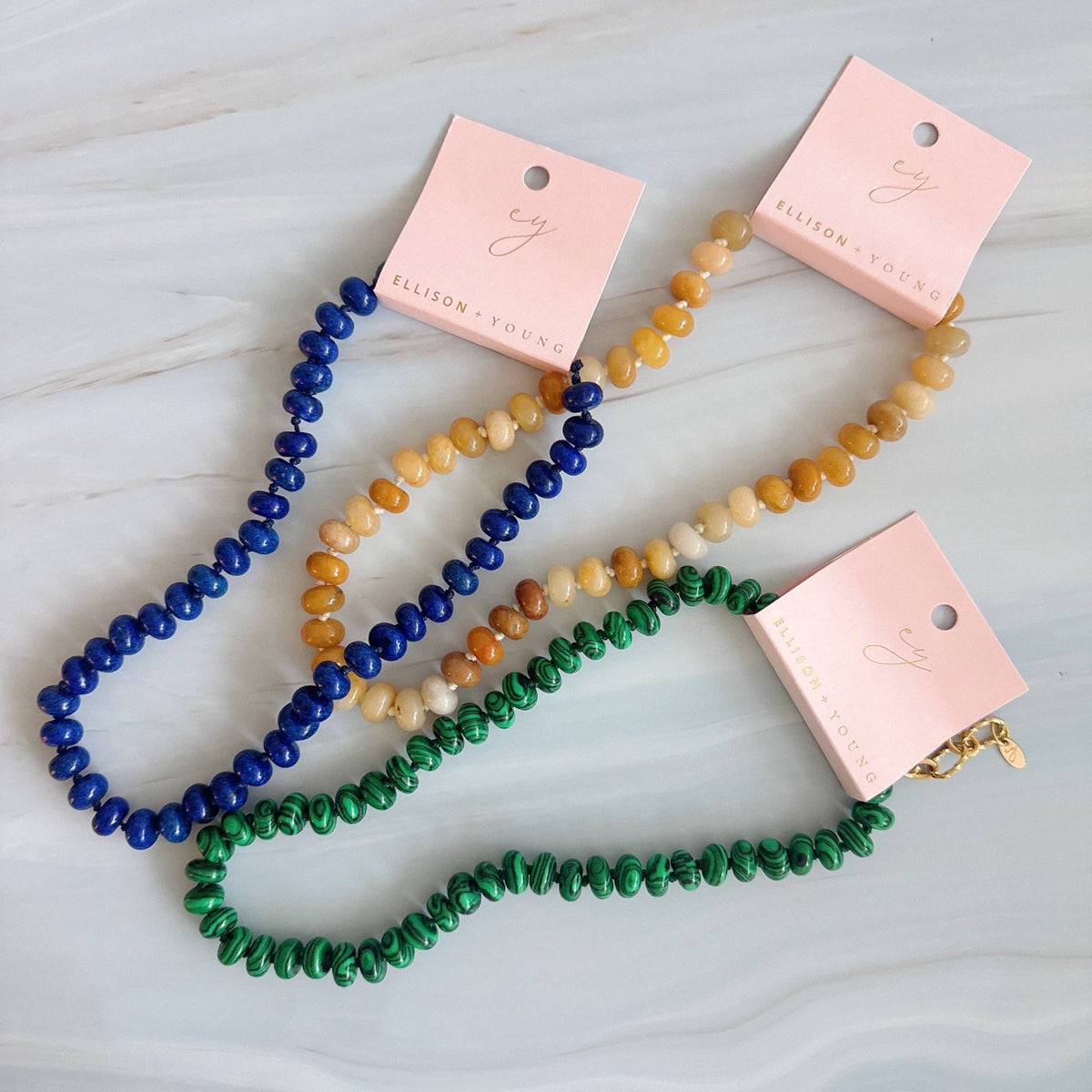 Three beaded necklaces in blue, orange, and green on pink cards with 'Elyse' branding.