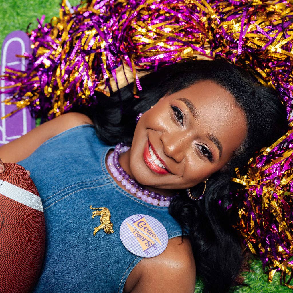 A smiling woman lying on artificial turf with purple and gold pom-poms around her, wearing a denim top and a round pin that reads 'Queen Chix'.