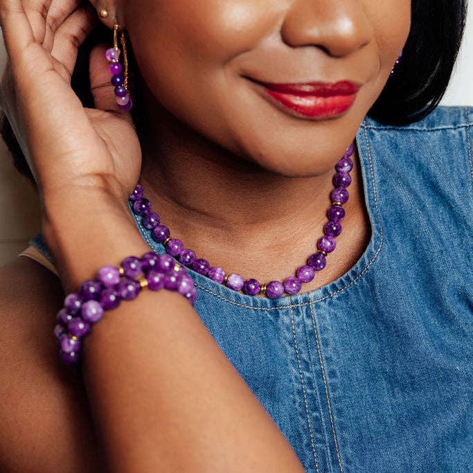 A close-up lifestyle shot of a woman wearing a matching purple beaded necklace and bracelet set with gold hoop earrings, paired with a blue denim top.