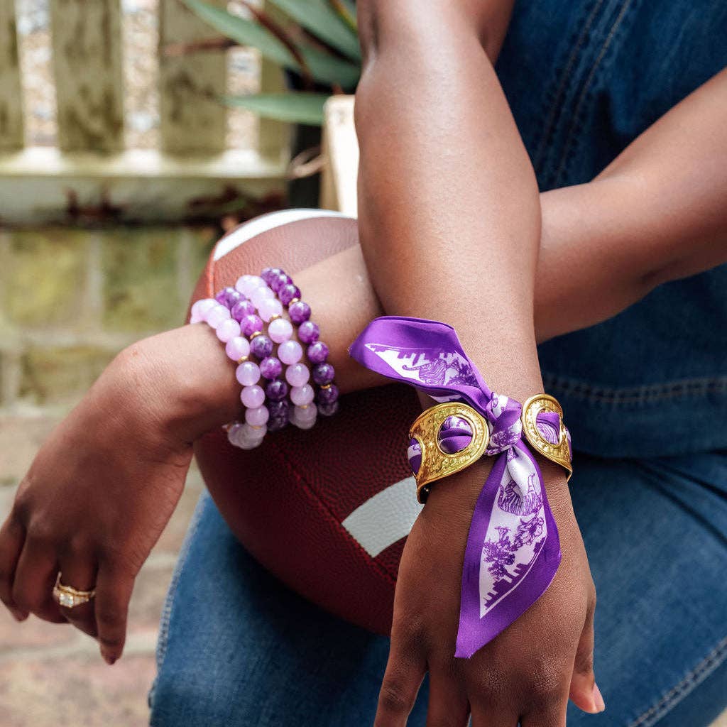 A close-up lifestyle shot of a person's arms and hands wearing multiple bracelets. The most prominent product is a purple and white patterned silk ribbon bracelet with gold-toned metal accents, styled on a person wearing blue jeans.