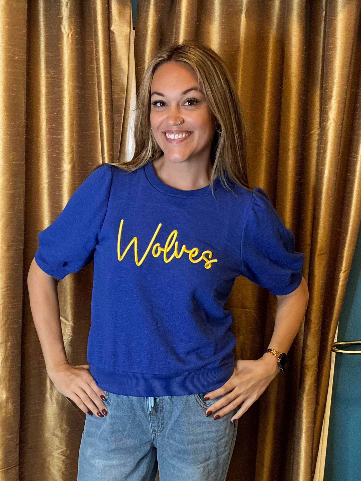 A woman smiling and posing in front of gold curtains, wearing a royal blue crewneck sweatshirt with 'Wolves' in puff embroidery  yellow script across the chest, paired with light blue jeans.