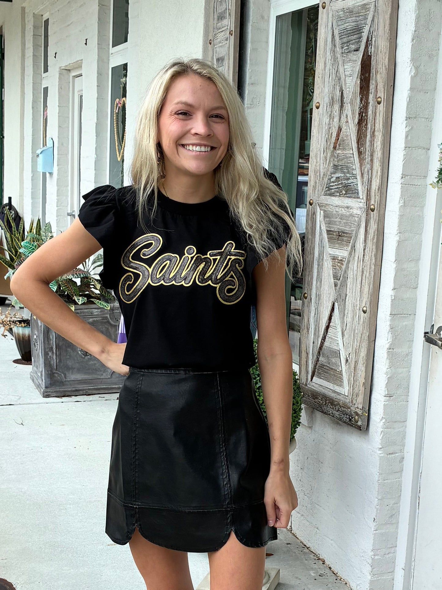 A woman stands on a storefront sidewalk wearing a black short-sleeve t-shirt with 'Saints' in black and gold script and a black skirt. 