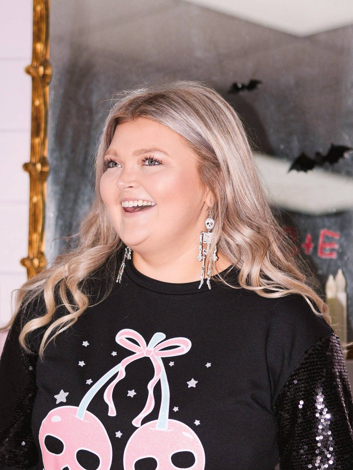 A woman wearing a paired of silver dangling skeleton earrings, set against a Halloween-themed backdrop.