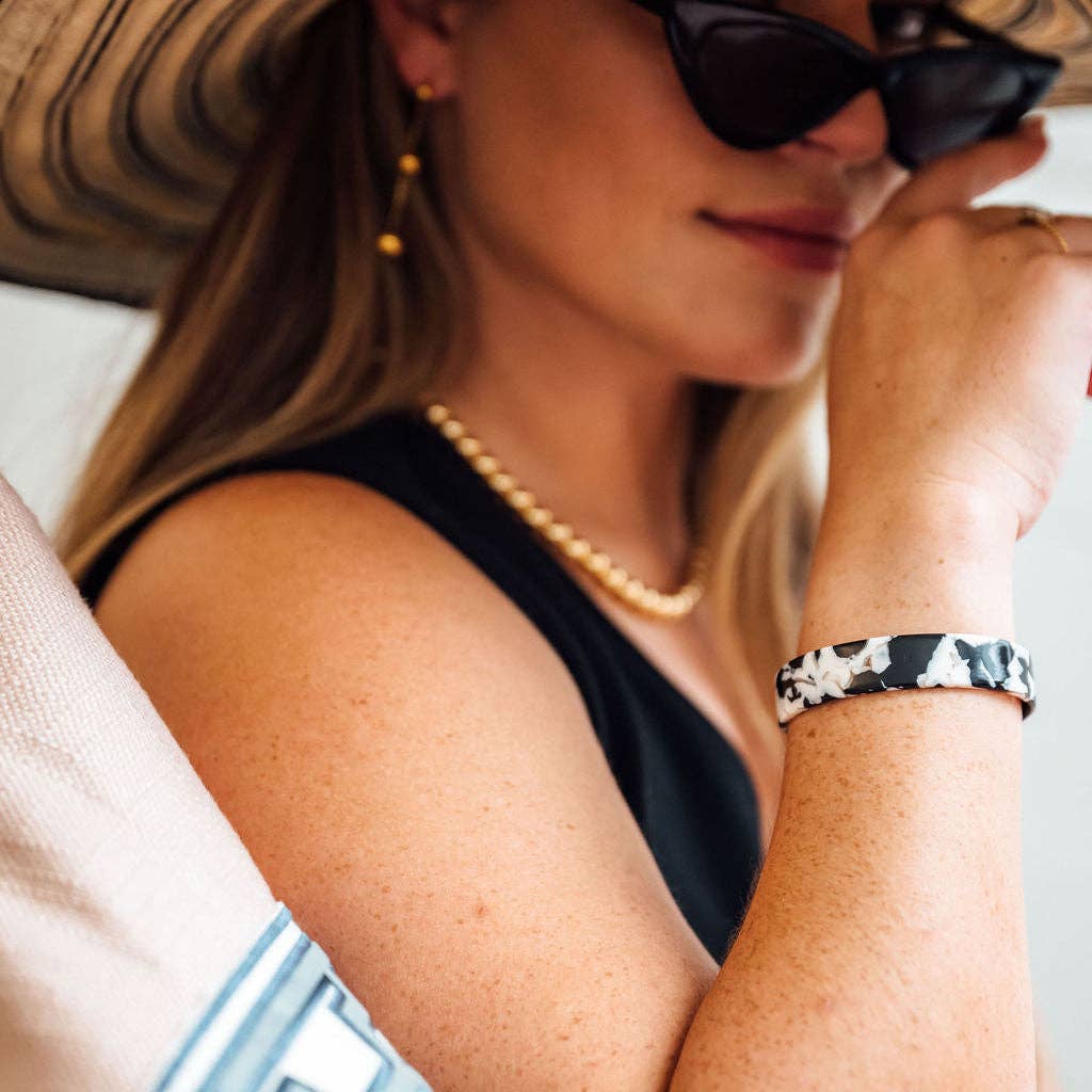 A close-up lifestyle shot of a woman wearing a black and white patterned silicone bracelet on her wrist, along with a pearl necklace, gold hoop earrings, and a black top.