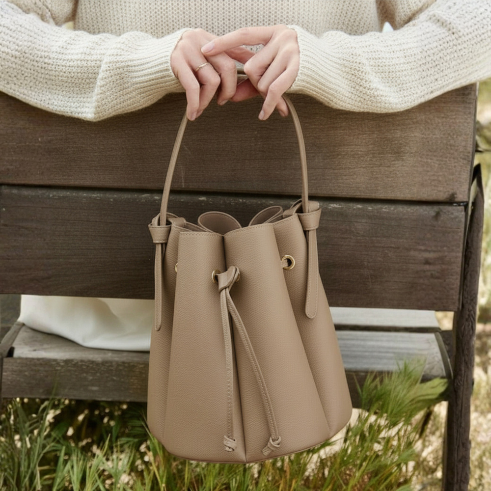 Beige leather bucket bag held by a person wearing a cream sweater on a wooden bench.