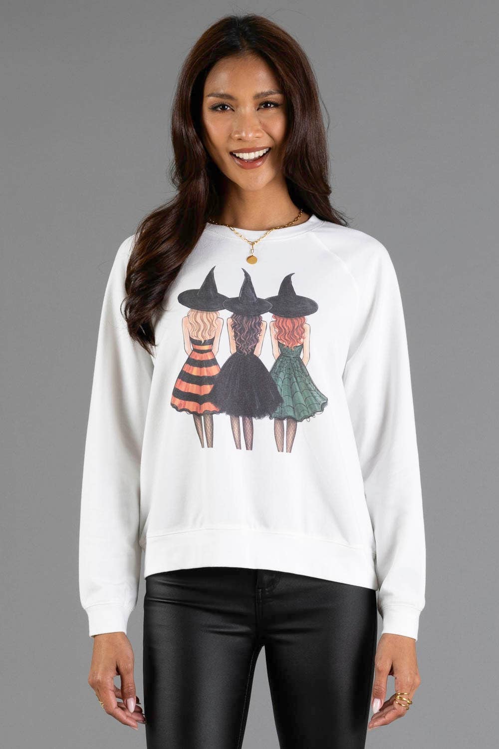 A woman wearing a white crewneck sweatshirt with a graphic of three girls dressed as witches, paired with black pants, against a solid gray studio background.