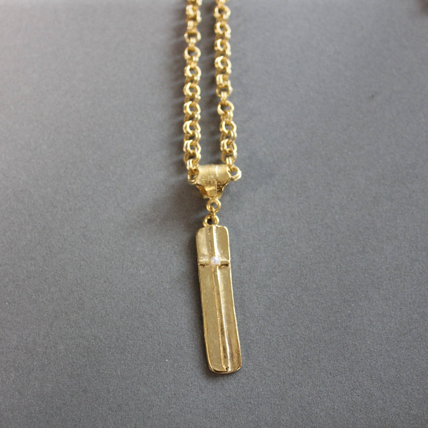 A gold necklace with a polished rectangular bar pendant featuring a raised cross detail, displayed on a neutral gray background.