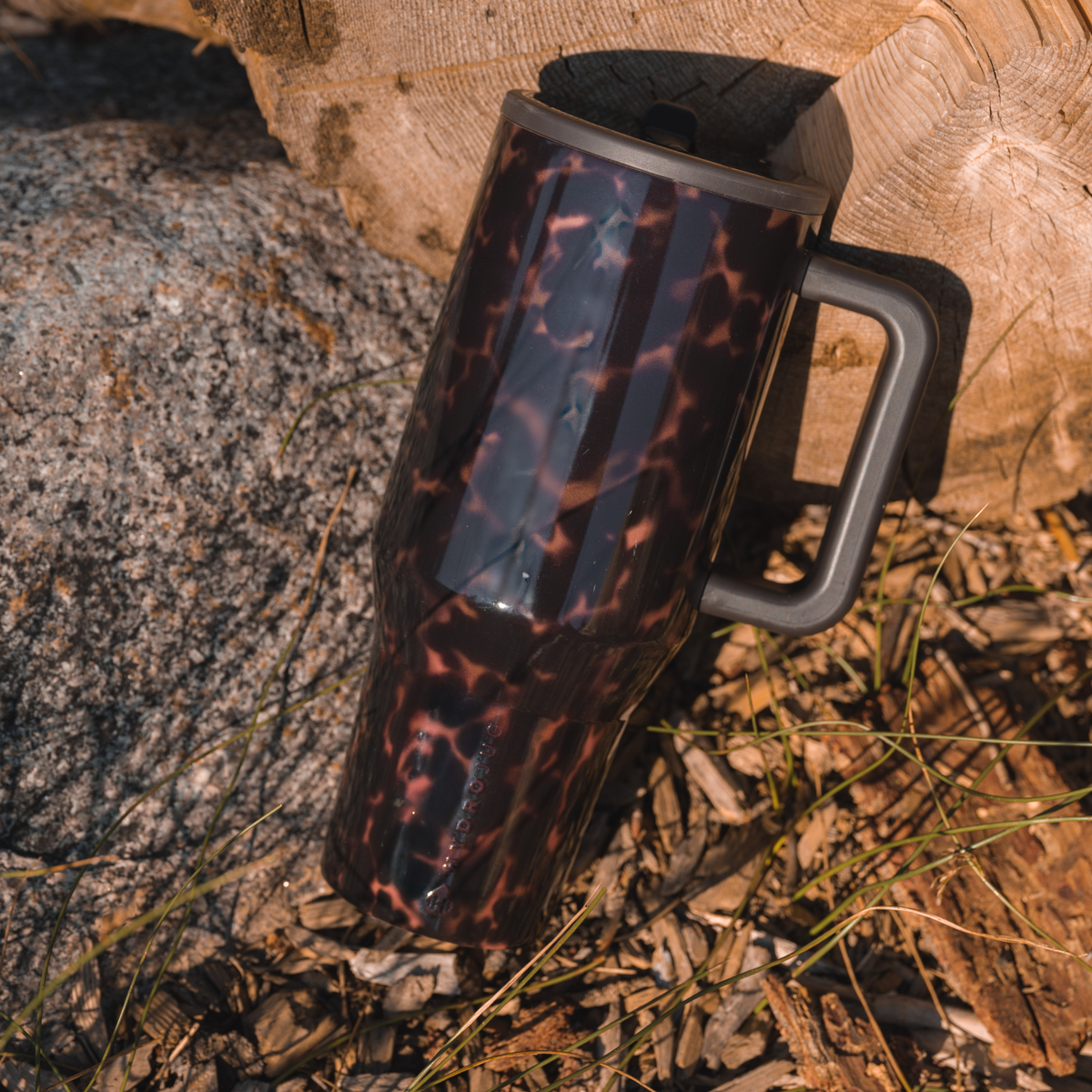Mocha Tortoise HydroJug tumbler resting against cut logs outdoors, showing its glossy brown tortoiseshell pattern