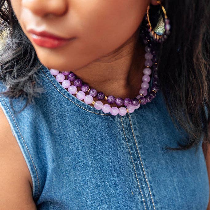 A close-up lifestyle shot of a woman wearing a double-strand beaded necklace and matching drop earrings. The beads are a mix of purple amethyst and pale pink quartz, separated by small gold spacer beads, and worn with a blue denim top.