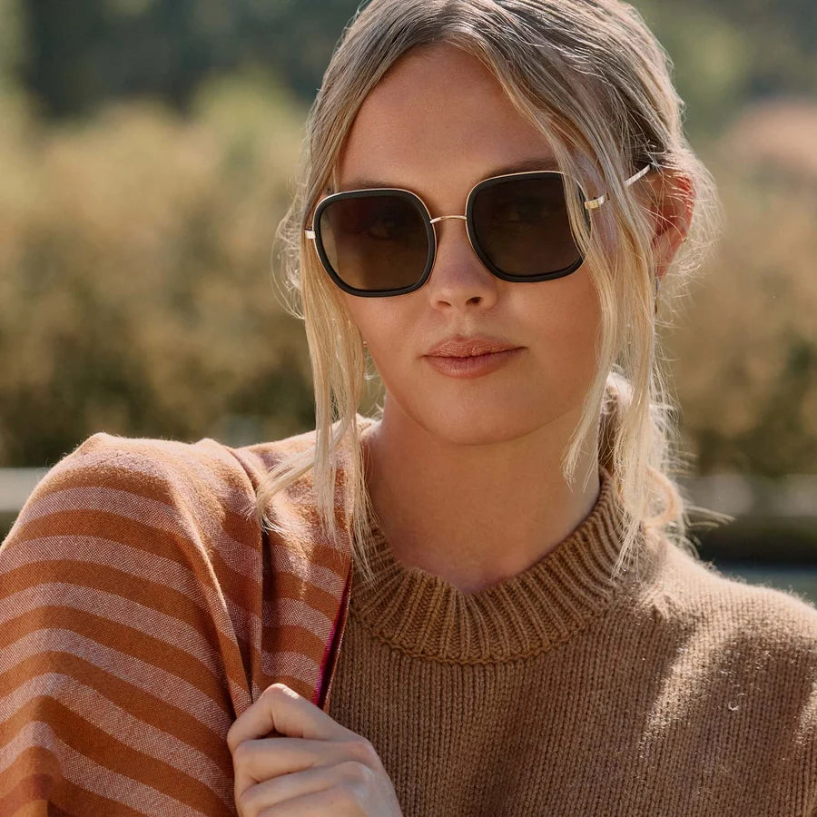 A woman wearing square dark sunglasses with thin gold metal frames and a brown ribbed knit sweater, photographed outdoors with a blurred natural background.