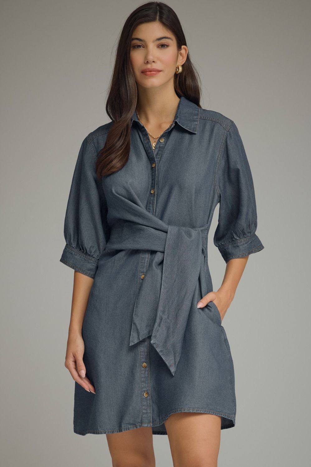 A woman modeling a slate blue denim shirt dress with puff sleeves, a button-down front, and a self-tie belt, set against a solid gray studio background.