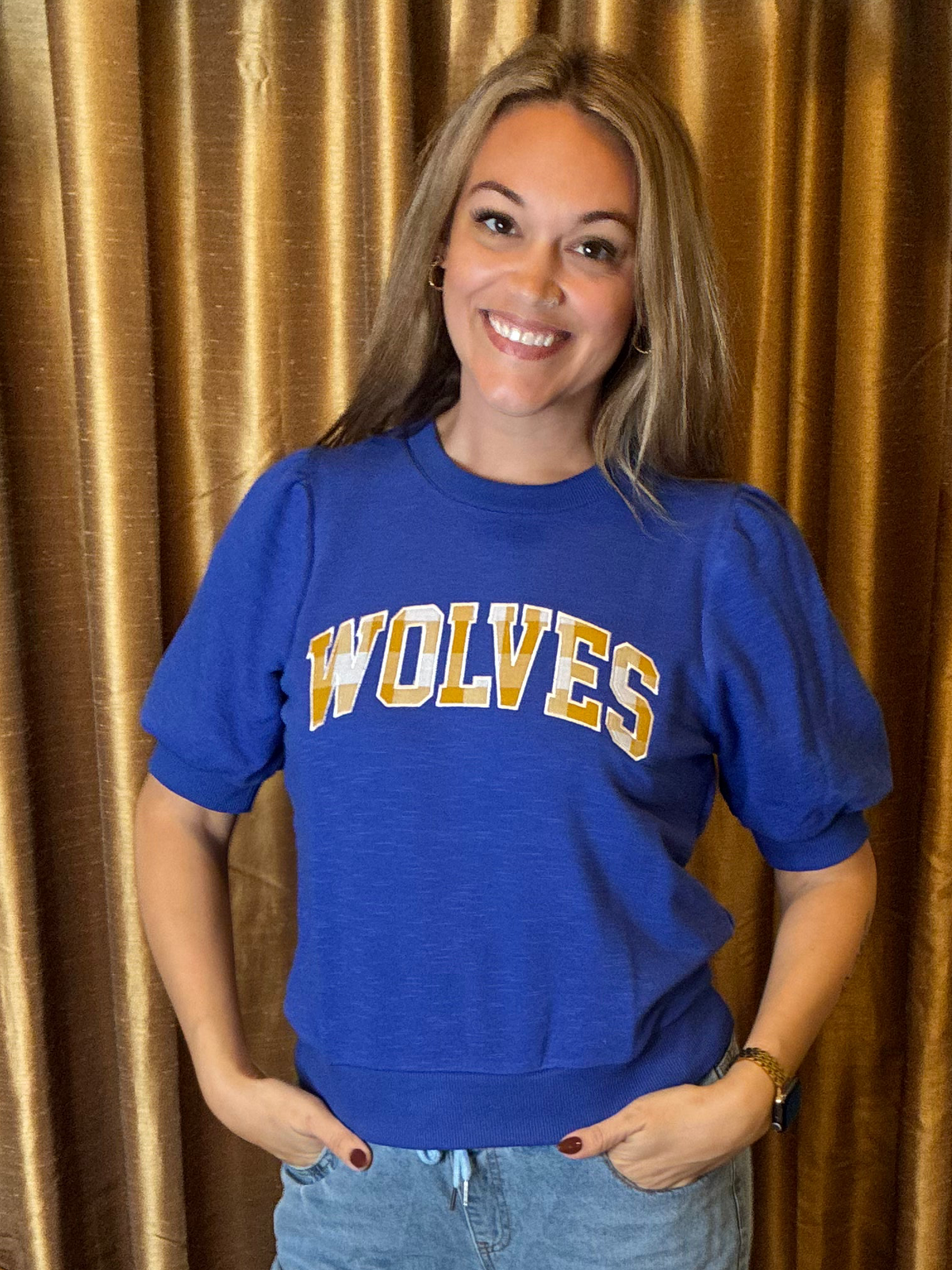 A smiling woman with blonde hair stands in front of a gold curtain backdrop, wearing a royal blue short-sleeve crewneck sweatshirt with 'WOLVES'  across the chest in yellow and white varsity lettering, paired with light blue jeans.