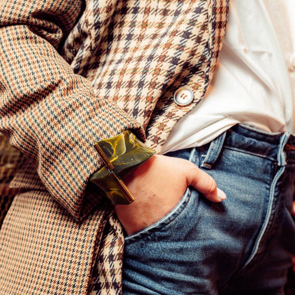 A close-up lifestyle shot of a person wearing a chunky, wide rectangular tortoiseshell-style bangle with a gold-toned cross accent on the back, styled with a brown and beige houndstooth blazer, white shirt, and blue jeans.