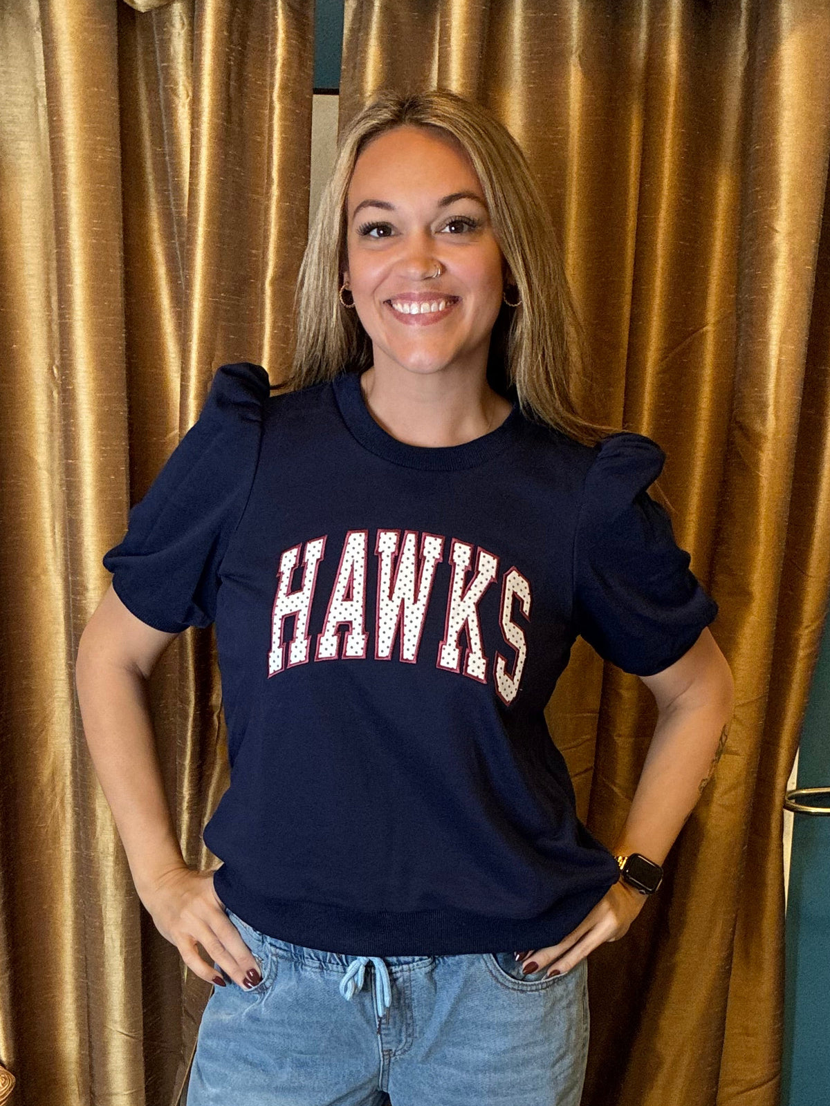 A woman stands indoors wearing a navy blue short-sleeve graphic T-shirt with 'HAWKS' in white block letters with polka dots across the chest, paired with light-wash ripped jeans and black sandals. She is posed in front of gold curtains and a blue wall.