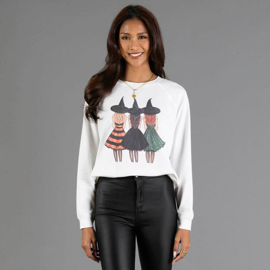 A woman modeling a white long-sleeve crewneck sweatshirt with a graphic of three girls in witch hats, paired with black pants, against a solid gray studio background.