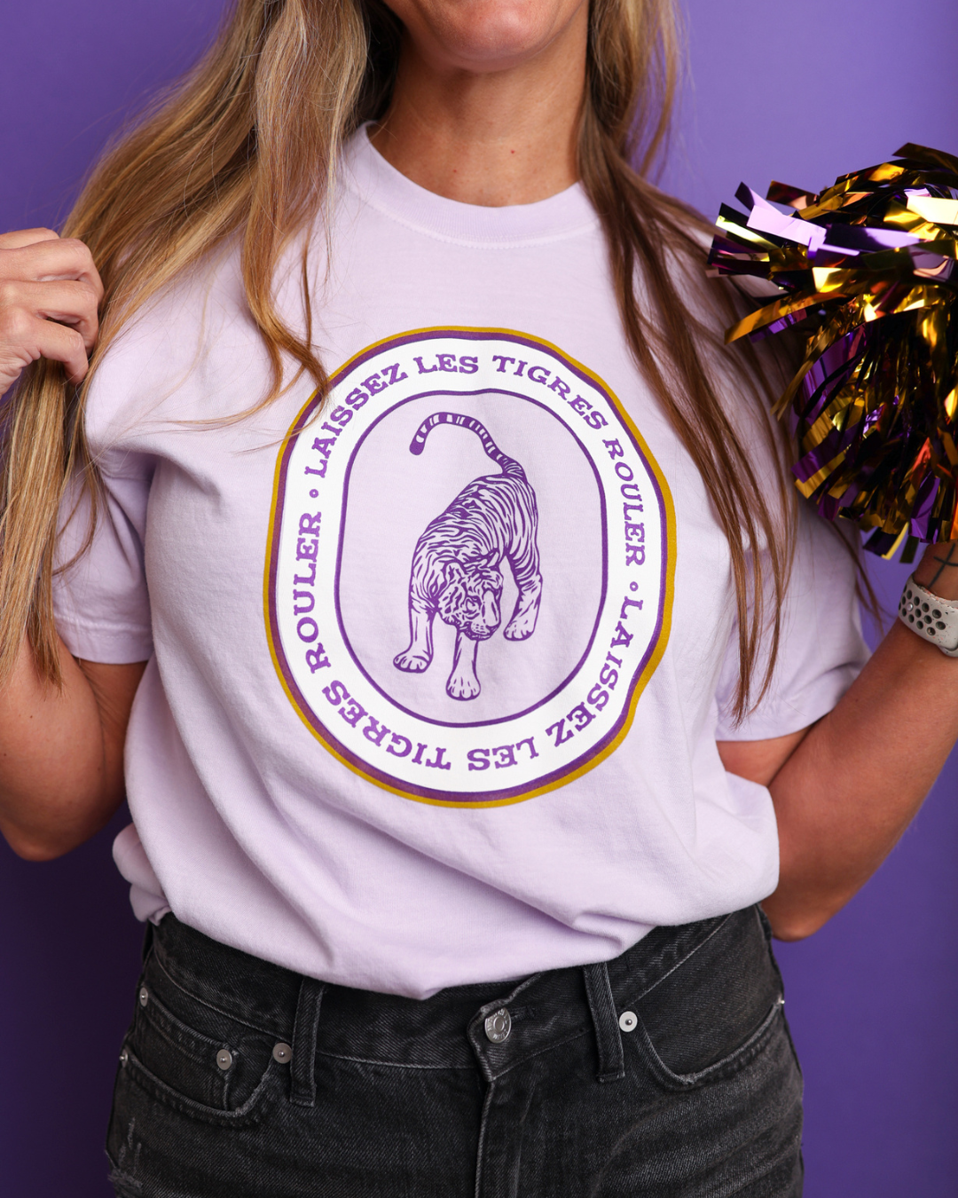 A woman wearing a lavender graphic t-shirt with a purple and gold tiger design and French text, paired with dark gray jeans, holding a purple and gold cheer pom-pom against a solid purple background.