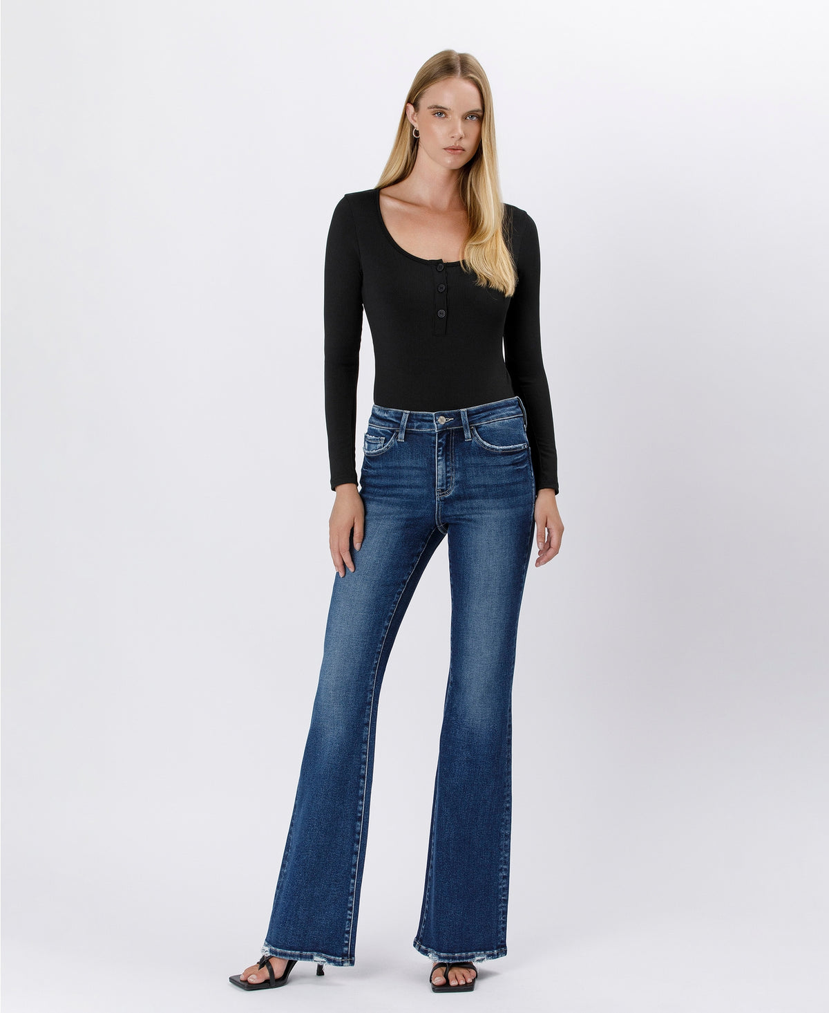 A model stands against a light gray studio background wearing a fitted black long-sleeve top with a scoop neckline and blue flare jeans, paired with black sandals.