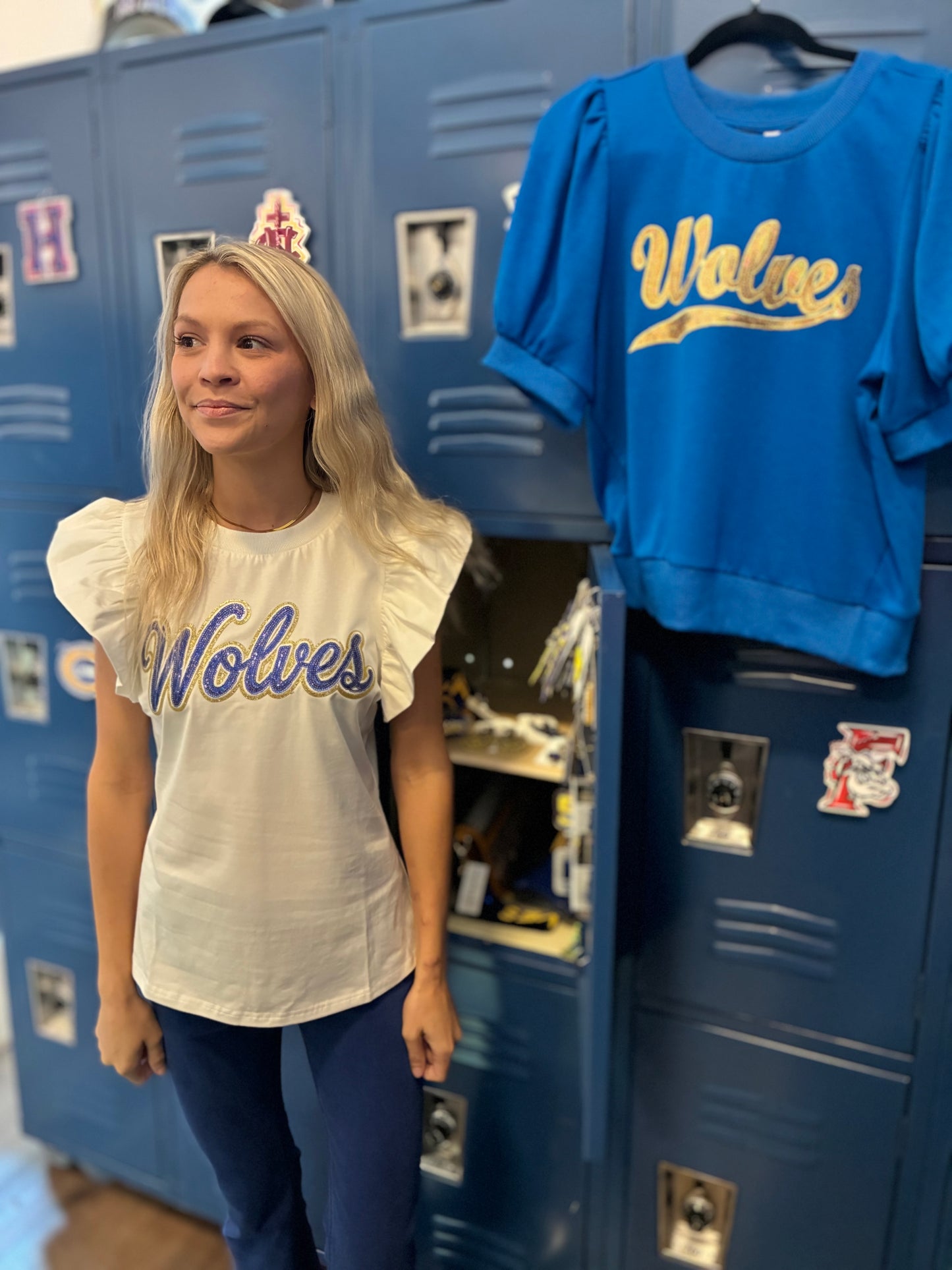 A woman in a clothing store in front of lockers wearing a blue and gold Wolves flutter sleeve top with other Wolves items in the background.