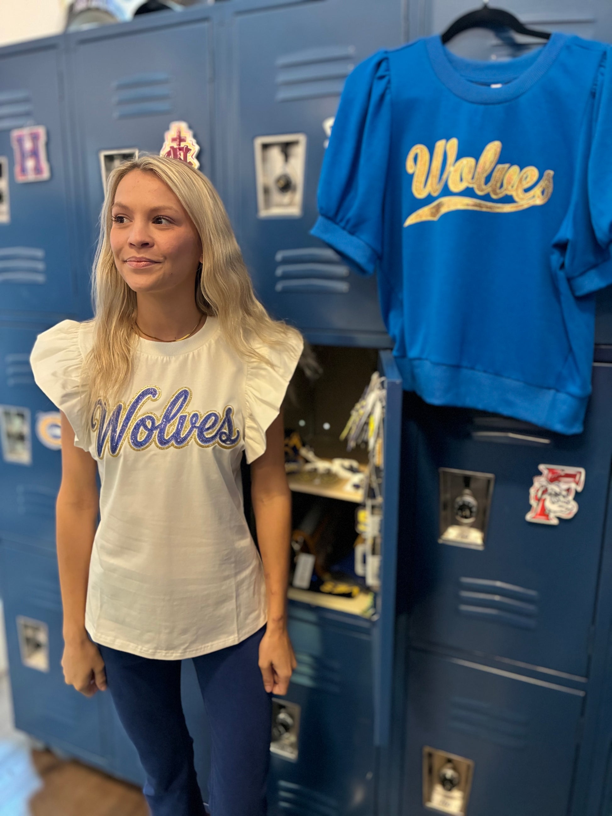 A woman in a clothing store in front of lockers wearing a blue and gold Wolves flutter sleeve top with other Wolves items in the background.