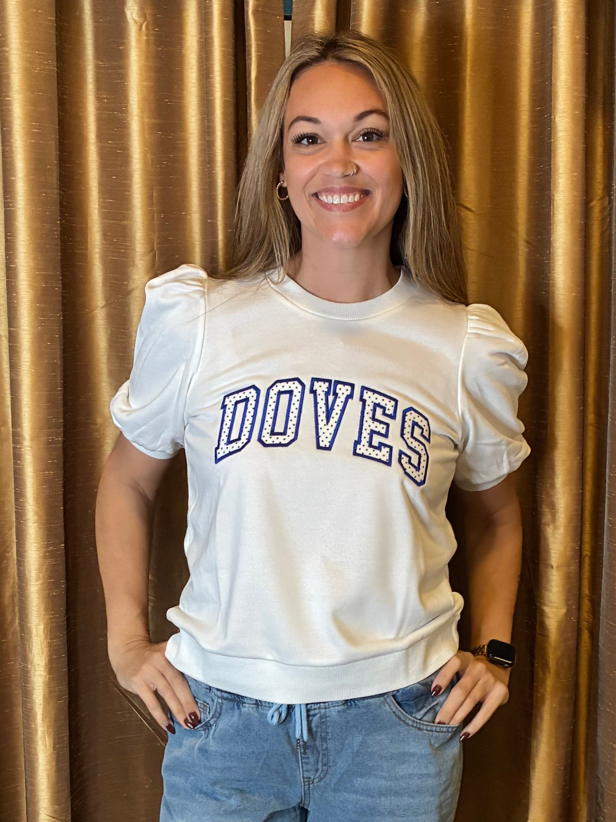 A woman stands in front of gold curtains wearing a white puff-sleeve sweatshirt with 'DOVES' embroidery appliquéd , paired with light-wash ripped jeans and black sandals.