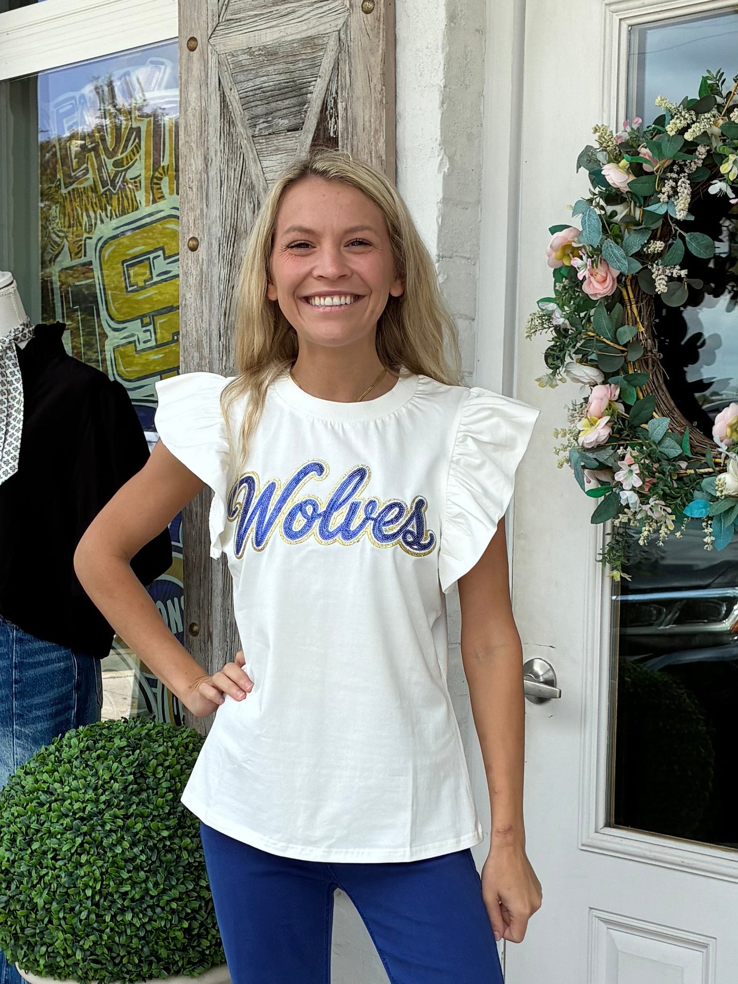 A woman stands on a storefront porch wearing a white ruffle-sleeve t-shirt with 'Wolves' printed in blue script, paired with blue flared pants and silver sneakers.