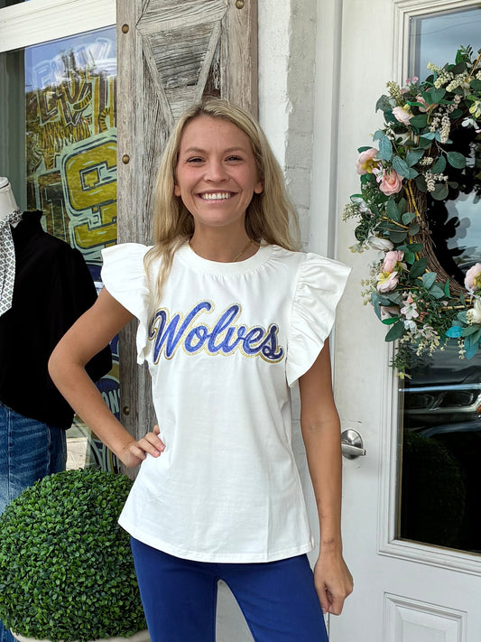 A woman stands on a storefront porch wearing a white ruffle-sleeve t-shirt with 'Wolves' printed in blue script, paired with blue flared pants and silver sneakers.