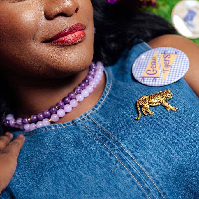 A close-up lifestyle shot of a woman wearing a gold tiger-shaped lapel pin on a blue denim top, alongside a purple beaded necklace and a round 'Cactus Tigers' brand patch.