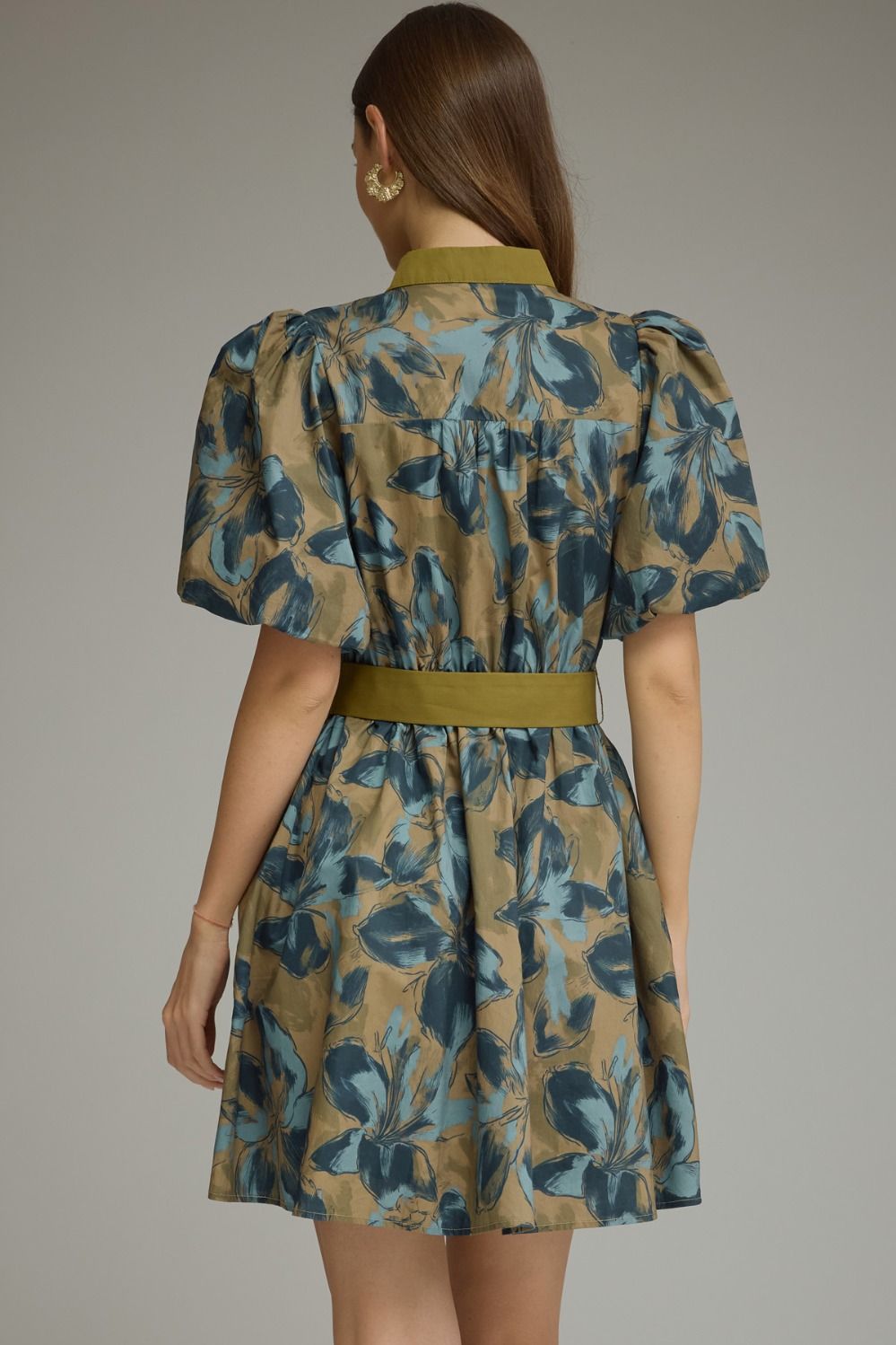 A woman shown from the back wearing a short puff-sleeve dress with a blue and teal floral print on a tan background, featuring an olive green collar and waist belt, set against a plain gray studio background.