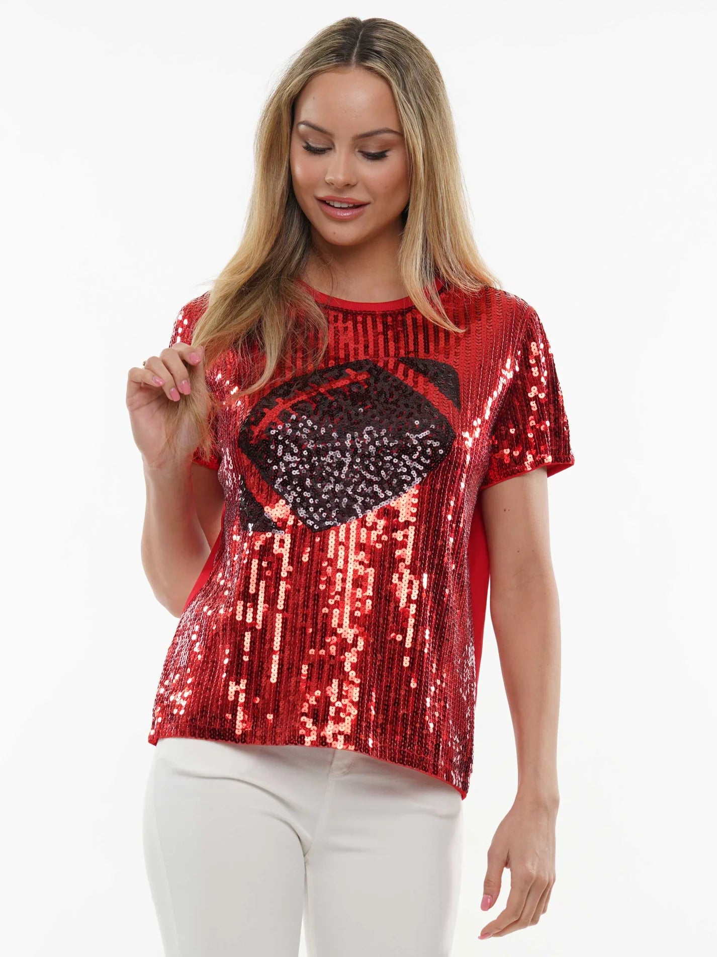 A woman wearing a red sequined short-sleeve top with a large black sequined graphic on the front, paired with white pants, against a plain white background.