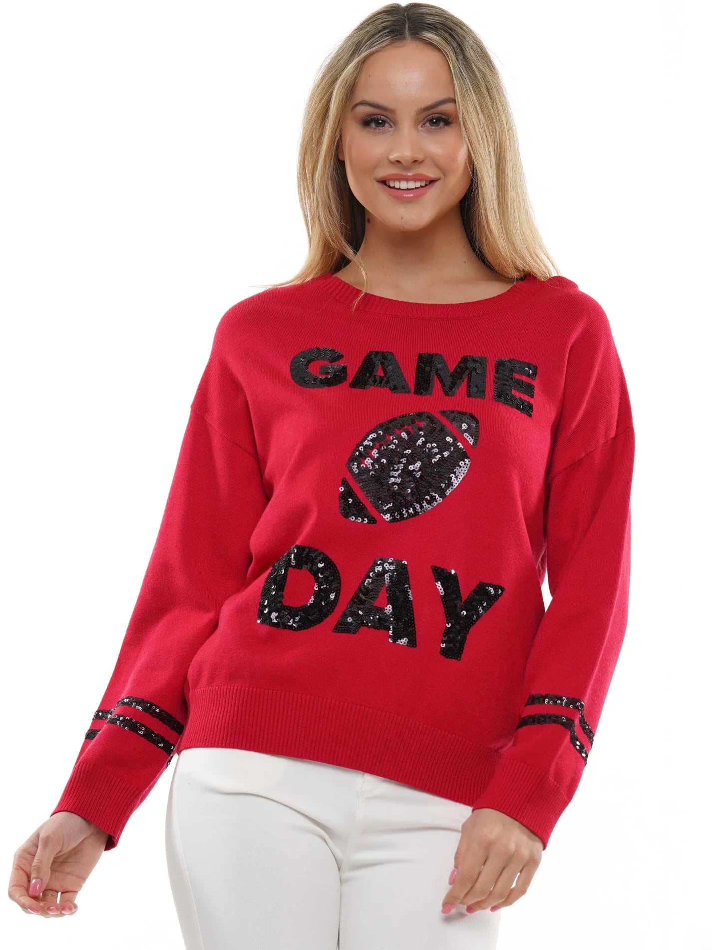 A woman wearing a bright red crewneck sweater with black sequined 'GAME DAY' lettering and a football graphic on the front, paired with white pants, against a plain white background.