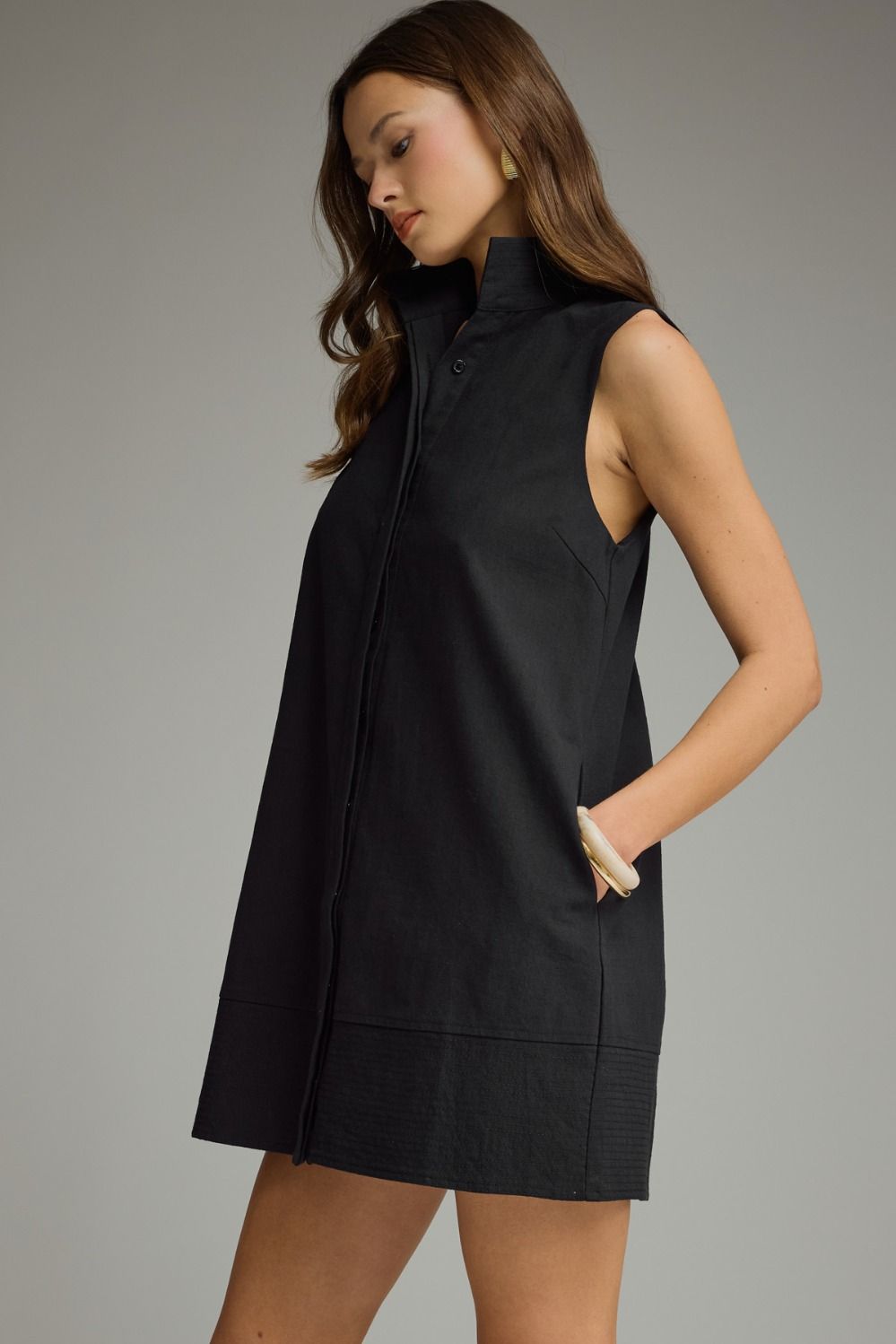 A woman wearing a sleeveless black mini dress with a high mock neck and front button placket, posed against a neutral gray studio background.