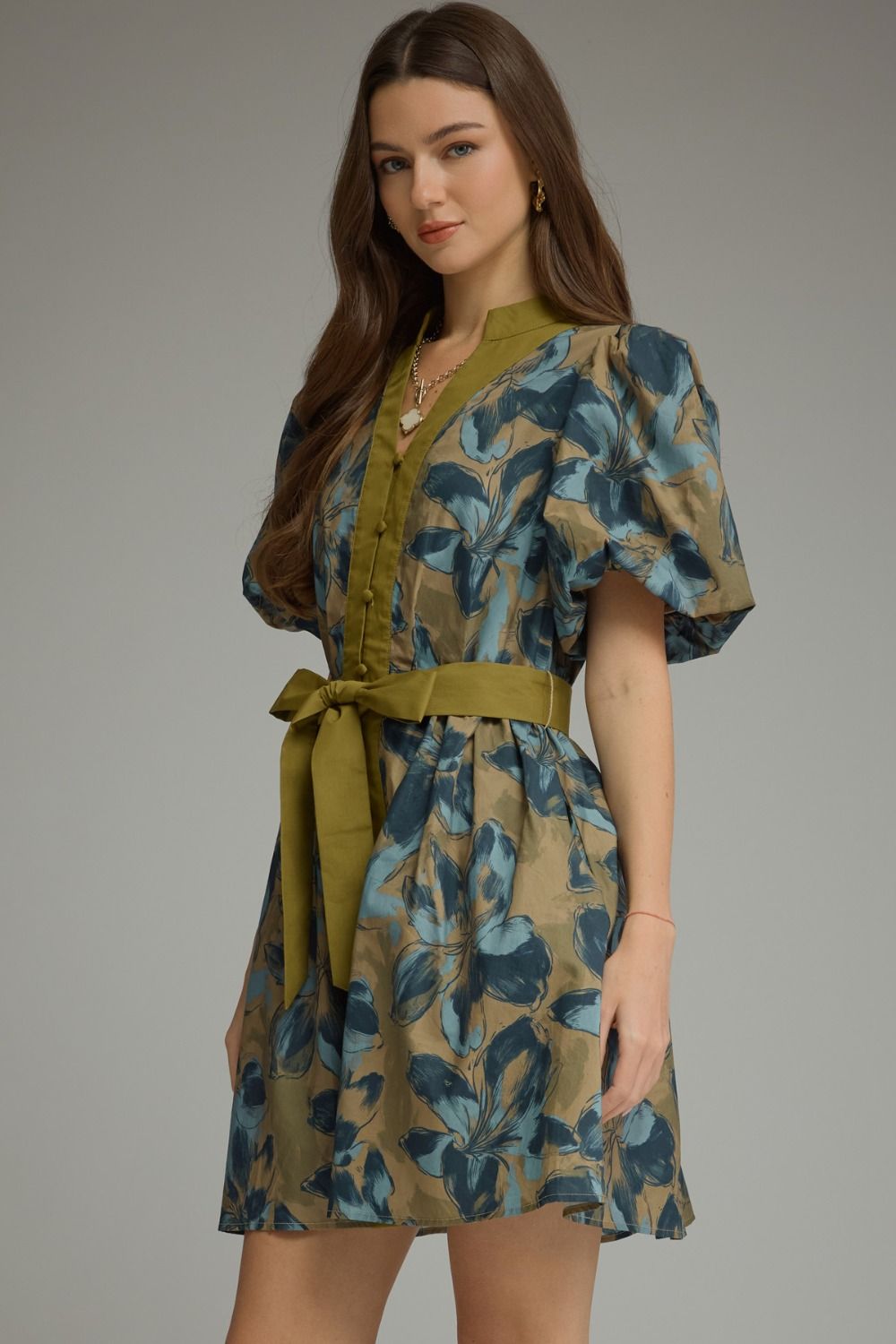 A woman modeling a short puff-sleeve dress with a blue and green leaf pattern on a tan base, featuring an olive green waist tie and collar, set against a plain gray studio background.