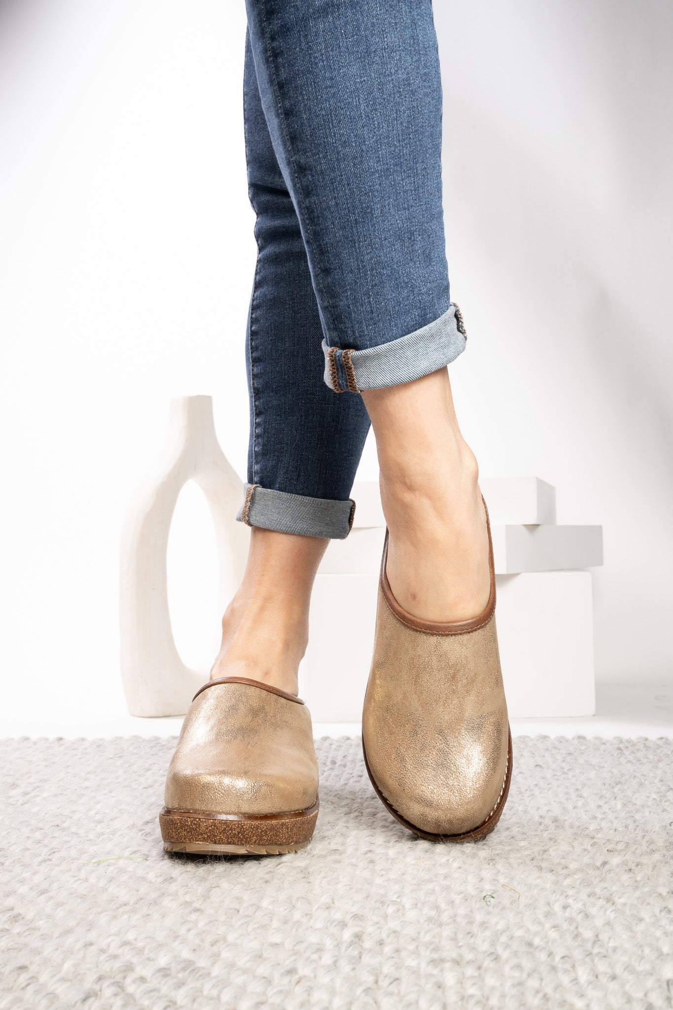 A person wearing blue cuffed jeans and a pair of distressed metallic gold slip-on mules on a light gray textured rug, with a white background and minimalist white decor.